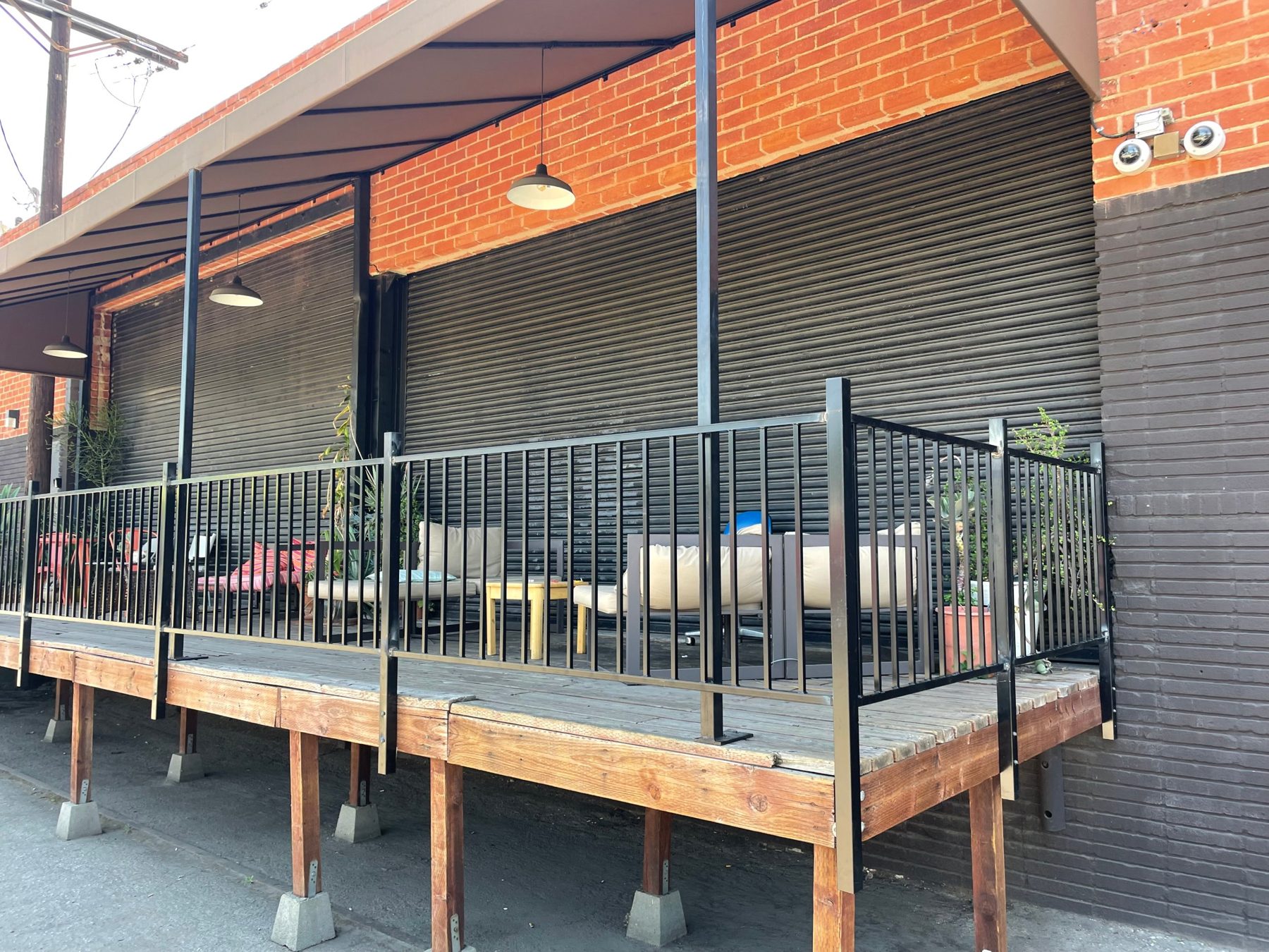 Covered, railed wooden deck with lounge seating, plants and pendant lights against the brick building